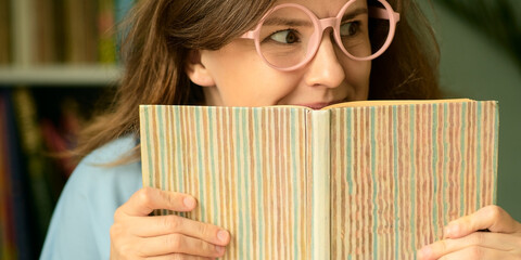 A young woman wearing pink round glasses sits at a table peeking from behind an interesting book. She looks sideways with a joyful and sly expression, anticipating reading.