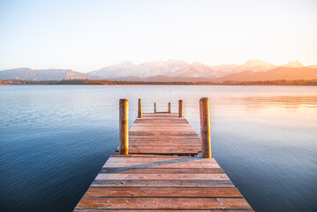 View of a wooden pier stretching into the serene lake, with snow-capped mountains glowing under the soft light, Hopfen am See, Fussen, Bavaria, Germany.