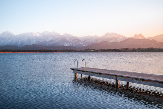 View of a serene lake reflecting the soft hues of dawn, framed by snow-capped mountains and a rustic pier, creating a tranquil scene, Fussen, Bavaria, Germany.