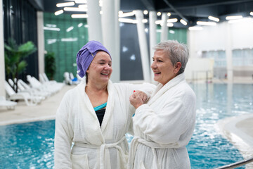 Two cheerful senior women in white bathrobes laughing and relaxing together at an indoor swimming...