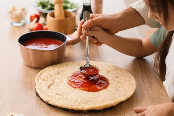 Father with his daughter applying tomato sauce on pizza dough in kitchen, closeup