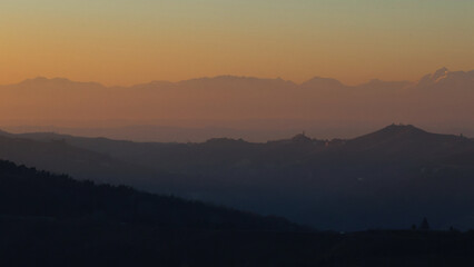 Stunning evening sunset over Piedmont's Langhe and Asti region, with the majestic snow-capped Monte Rosa and Alps glowing in warm alpenglow against a dramatic pink-orange sky. Perfect panoramic vista 