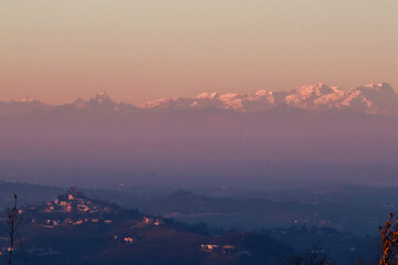 Stunning evening sunset over Piedmont's Langhe and Asti region, with the majestic snow-capped Monte Rosa and Alps glowing in warm alpenglow against a dramatic pink-orange sky. Perfect panoramic vista 