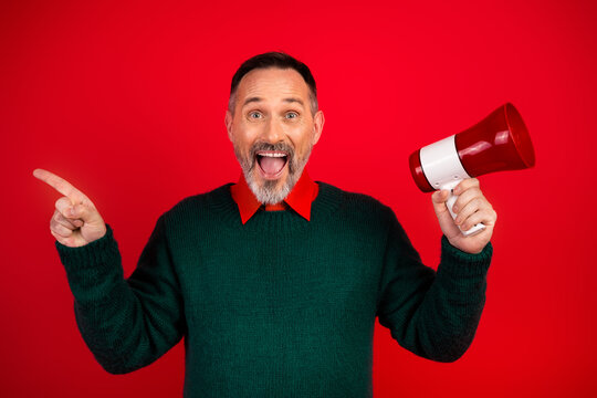 Funny mature man in green sweater shouts cheer with megaphone Christmas promotion on bright red background during festive season