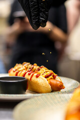 A close-up vertical shot of a person in black gloves sprinkling toppings onto a gourmet hot dog with ketchup, mustard, bacon bits, and cheese in a bun, served on a plate.