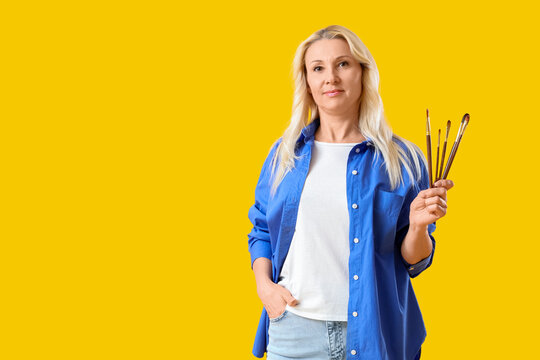 Mature blonde woman with paint brushes on yellow background
