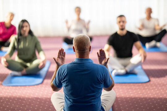 Rear view of a male instructor guiding a mixed age group during a stretching or rehabilitation class indoors. Participants are sitting on yoga mats following his movements. Concept of health, wellness