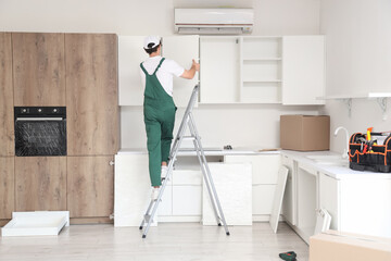 Male worker assembling cupboard on stepladder in kitchen, back view