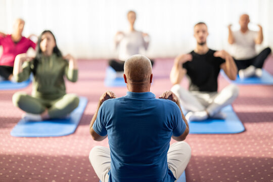 Rear view of a male instructor guiding a mixed age group during a stretching or rehabilitation class indoors. Participants are sitting on yoga mats following his movements. Concept of health, wellness