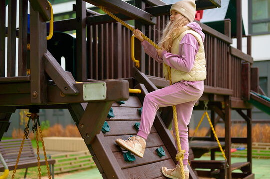 girl climbing playground structure wearing beige vest and pink tracksuit. outdoor adventure and active lifestyle for children. engages in play, balance, confidence building