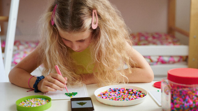 girl focused on arts and crafts using colorful beads with smartphone nearby. creative indoor hobby activity, promoting fine motor skills and concentration in playful environment - Powered by Adobe