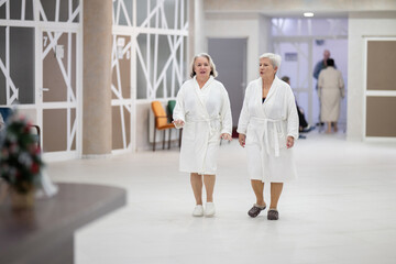 Elderly women walking together in a wellness and spa center dressed in white bathrobes, symbolizing relaxation, friendship, and healthy lifestyle.