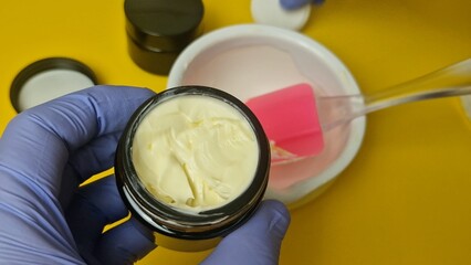 Extreme close-up view of a cosmetic chemist mixing a luxurious, yellow cream base in a white ceramic mortar with a pestle. Hands wear protective blue gloves. Perfect for beauty, pharmacy, and skincare
