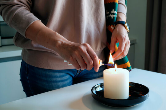 Young adult woman lighting large candle with lighter on black plate, hands and torso visible, wearing casual clothing, standing at table in modern indoor setting - Powered by Adobe