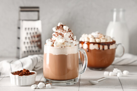 Glass cups of hot cocoa drink with whipped cream and marshmallows on white tile table