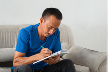 Focused Latino man writing in notebook on his lap, seated on couch, left hand missing four fingers evident