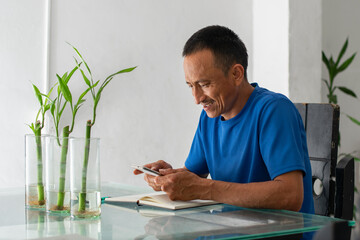 Smiling Latino man at a table using a smartphone, informal home scene, with four fingers amputated on his left hand.