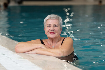 Smiling senior woman enjoying relaxation in an indoor swimming pool, leaning on the poolside. Concept of active lifestyle, wellness, health, hydrotherapy, and leisure for older adults.