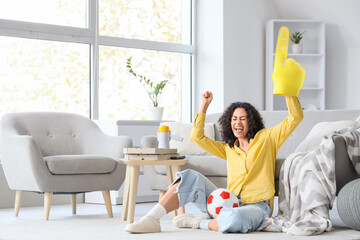 Female African-American sports fan with foam hand and soccer ball at home