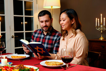 Caucasian man and Asian woman sitting at dinner table reading Jewish prayer book together, celebrating traditional holiday meal with wine, candles and festive dishes visible on table