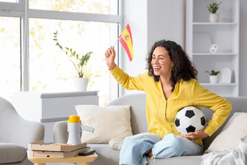 Female African-American sports fan with Spanish flag and soccer ball on sofa at home