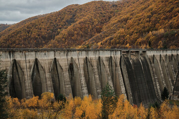 Scenic view of a dam in the mountains during autumn. The grey concrete structure contrasts with the colorful foliage, creating a powerful image of nature and engineering.