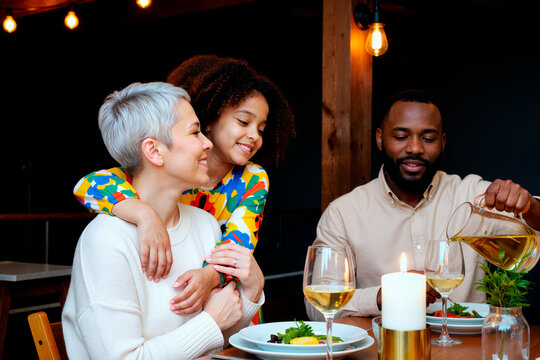 Caucasian middle aged woman smiling while being hugged by Black child girl, Black man pouring drink at dinner table, family sharing meal together in modern dining setting - Powered by Adobe