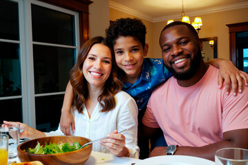 Caucasian woman, Black man, and teenage boy smiling together at dining table, teenage boy standing behind adults with arms around their shoulders, sharing family meal