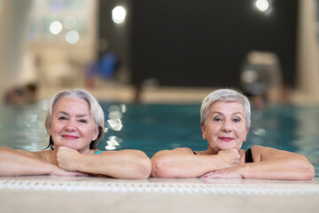 Two smiling senior women enjoying leisure time in an indoor swimming pool, leaning on the poolside. Concept of friendship, wellness, active lifestyle, relaxation, and healthy aging.