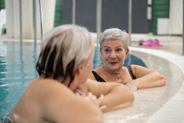 Two smiling senior women enjoying leisure time in an indoor swimming pool, leaning on the poolside. Concept of friendship, wellness, active lifestyle, relaxation, and healthy aging.