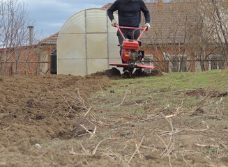 A farmer in dark clothes plows the land on his personal plot with a red iron petrol cultivator, a man loosens the soil in a village using a mechanized plow against the background of a brick house