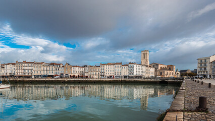 A panoramic view of La Rochelle's Vieux Port, featuring the iconic medieval defense towers and historic white buildings reflected in the still harbor water. Numerous yachts are docked under a dramatic