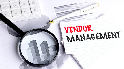 Vendor management text on note card with magnifying glass, pen and financial graphs on desk