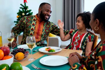 Black man smiling and giving high five to Black girl while sitting at dining table with Black woman, celebrating festive occasion with food and drinks, Christmas tree in background