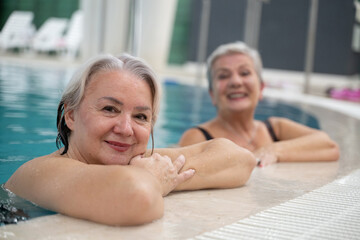 Two smiling senior women enjoying leisure time in an indoor swimming pool, leaning on the poolside. Concept of friendship, wellness, active lifestyle, relaxation, and healthy aging.