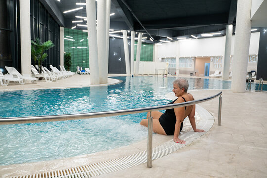 Elderly woman sitting by the edge of a modern indoor spa pool, enjoying the relaxing water jets. Wellness and relaxation concept in a luxurious spa or wellness center.