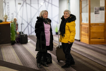 Two older women dressed in winter clothing standing in a hotel lobby and having a friendly...