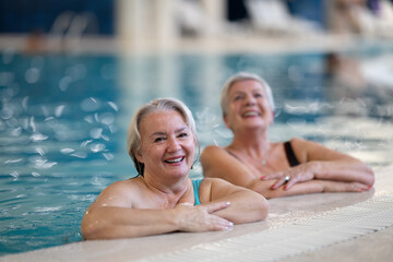 Two smiling senior women enjoying leisure time in an indoor swimming pool, leaning on the poolside. Concept of friendship, wellness, active lifestyle, relaxation, and healthy aging.