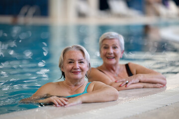 Two smiling senior women enjoying leisure time in an indoor swimming pool, leaning on the poolside. Concept of friendship, wellness, active lifestyle, relaxation, and healthy aging.