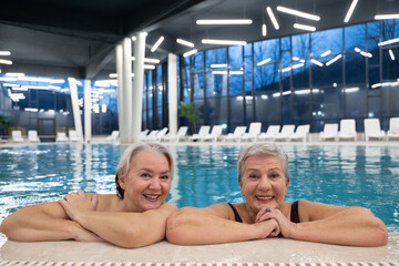 Two smiling senior women enjoying leisure time in an indoor swimming pool, leaning on the poolside. Concept of friendship, wellness, active lifestyle, relaxation, and healthy aging.