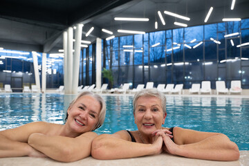 Two smiling senior women enjoying leisure time in an indoor swimming pool, leaning on the poolside. Concept of friendship, wellness, active lifestyle, relaxation, and healthy aging.