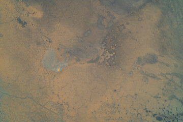 Aerial view of a dry landscape with cracked soil texture and patches of dried mud. The surface shows erosion patterns and natural fracturing typical of arid or drought-affected areas