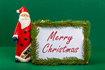 Merry Christmas. Santa Claus. Festive Santa Claus figurine in red attire stands next to decorative frame adorned with green foliage, displaying cheerful holiday greeting, creating joyful atmosphere