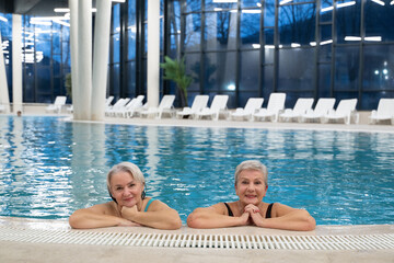 Two smiling senior women enjoying leisure time in an indoor swimming pool, leaning on the poolside. Concept of friendship, wellness, active lifestyle, relaxation, and healthy aging.