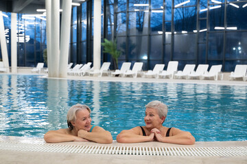 Two smiling senior women enjoying leisure time in an indoor swimming pool, leaning on the poolside. Concept of friendship, wellness, active lifestyle, relaxation, and healthy aging.