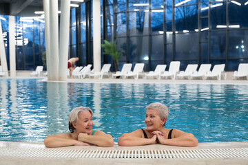 Two smiling senior women enjoying leisure time in an indoor swimming pool, leaning on the poolside. Concept of friendship, wellness, active lifestyle, relaxation, and healthy aging.