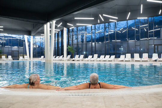 Two senior women enjoying relaxation while leaning on the edge of a modern indoor swimming pool in the evening. Concept of wellness, healthy lifestyle, spa, leisure, and friendship for older adults.