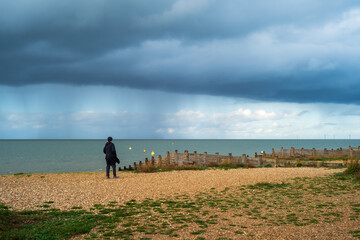 A pebble beach with wooden groynes in winter. There are rain clouds on the horizon. An unrecognisable man stands on the beach in a long coat and hat.
