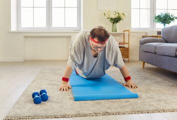 Overweight funny man doing pushups training at home. On mat, he attempts push-up with humor beside dumbbells. This fitness exercise supports better health. Playful perseverance in home sport workout.