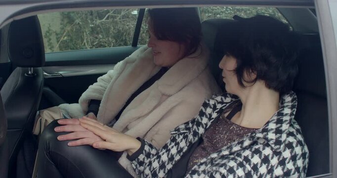 two women sitting in car, backseat pause during Prague bachelorette outing, muted winter coats, quiet conversation with hands resting on knees, candid friendship moment with urban foliage visible